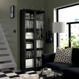 Bibliothèque BILLY noire avec livres et décoration par des escaliers blancs. Canapé sombre, guitare, fenêtre vue ville.