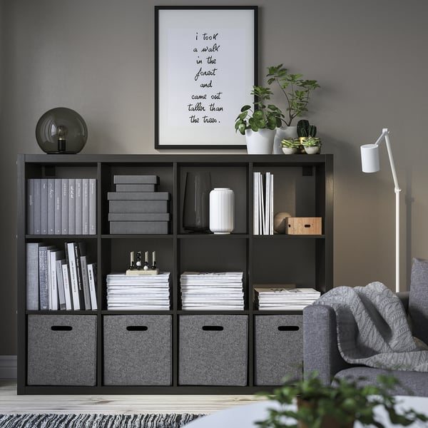 Étagère kallax noire avec boîtes de rangement en feutre BLÄDDRA, livres, plantes, œuvres d’art encadrées, lampe, canapé gris et tapis gris, organisés et soignés.