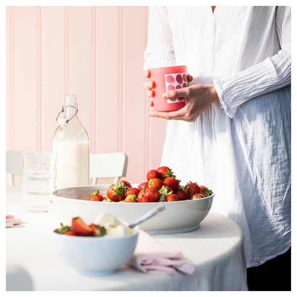 Personne tenant une tasse BLODBOK rouge avec des points près d’un bol de fraises sur une table rose clair.