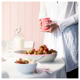 Personne tenant une tasse BLODBOK rouge avec des points près d’un bol de fraises sur une table rose clair.