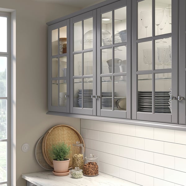 Armoire en verre BODBYN gris avec panneaux biseautés et poignées argentées, sur un comptoir en carrelage blanc contenant un plateau rond, une plante en pot et des bocaux.