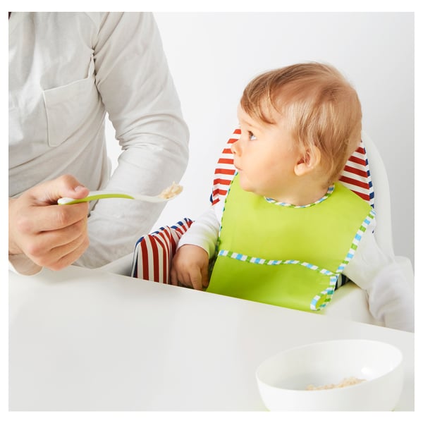 Une personne nourrissant un bébé avec une cuillère. Le bébé porte un bavoir vert avec une bordure rayée et lève les yeux attentivement. Un bol blanc est posé sur la table.
