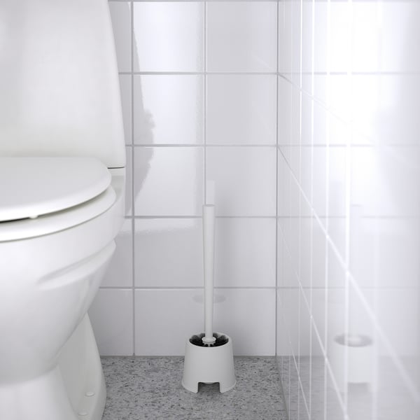 Une brosse de toilette blanche, BOLMEN, se trouve à côté d’une toilette sur un sol tacheté, prête à être nettoyée.