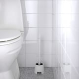 Une brosse de toilette blanche, BOLMEN, se trouve à côté d’une toilette sur un sol tacheté, prête à être nettoyée.