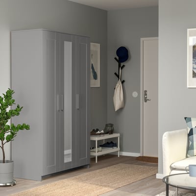 Entrée moderne avec armoire BRIMNES grise, porte-chaussures et porte-manteau. L’armoire reflète le mur, ajoutant de la profondeur. Décoré de plantes et d’objets.