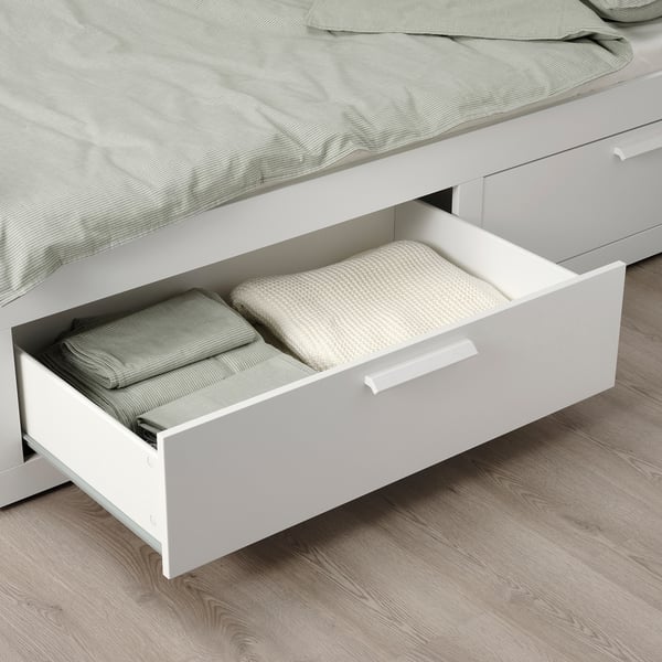 Un tiroir de sous-lit blanc avec des draps pliés, faisant partie d’un lit de jour BRIMNES peu encombrant avec rangement.