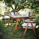 Ensemble de patio extérieur avec table et chaises en bois sous un arbre. La table a des assiettes, des tasses et des plantes en pot. Les chaises ont des coussins gris.