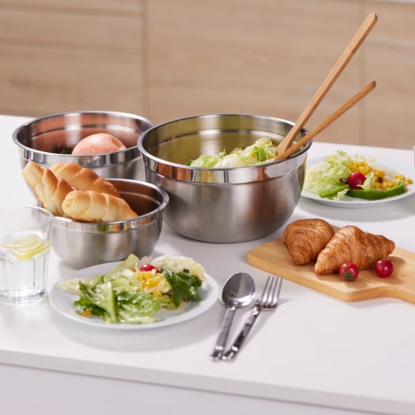 Trois bols à mélanger en acier inoxydable sur la table. Le bol du milieu contient de la salade avec des pinces. Assiettes, verre, couverts autour.