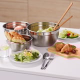 Trois bols à mélanger en acier inoxydable sur la table. Le bol du milieu contient de la salade avec des pinces. Assiettes, verre, couverts autour.