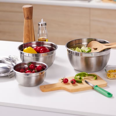 Bols en acier inoxydable avec couvercles pour la préparation, le service et le stockage des aliments. Nid de différentes tailles pour un rangement facile. Montré avec des légumes frais et une planche à découper.