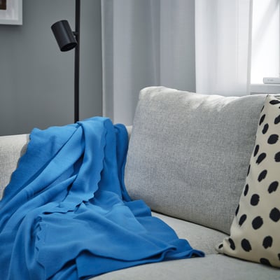 Un plaid en polaire DAGGKÅPOR bleu confortable, couvrant principalement un canapé gris clair, avec des coussins à pois noirs et une lampe à proximité.