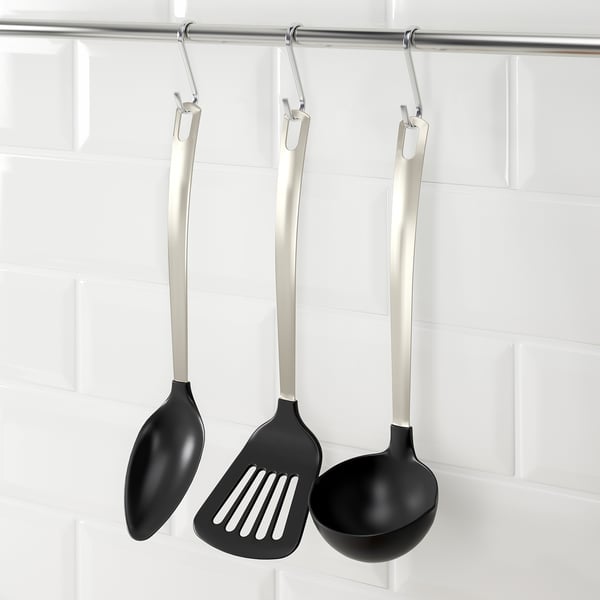 Trois ustensiles de cuisine noirs avec des poignées en argent sont suspendus à des crochets : cuillère, spatule et louche, conçus pour les casseroles et poêles antiadhésives.