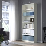 Étagère blanche avec des boîtes, des vases et des livres bleus. La chambre a une chaise, une fenêtre, une lampe.