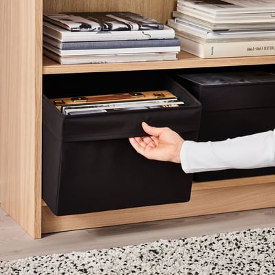 Une personne sort une boîte en tissu DRÖNA noir d’une étagère en bois, qui contient divers articles et est conçue pour maximiser l’espace de stockage.