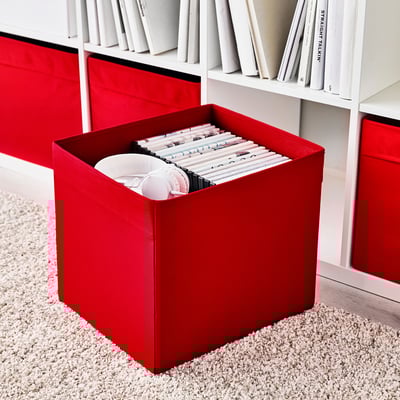 Boîte de rangement en tissu rouge, remplie d’objets, posée sur un tapis beige texturé. Partie de l’étagère kallax avec livres blancs.