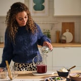 Une femme verse du sirop de myrtille d’une cruche transparente dans une autre cruche sur un comptoir de cuisine, entourée de verres et de nourriture.