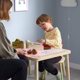 Enfant jouant avec la caisse enregistreuse de jouets DUKTIG à table avec de la nourriture de jeu. Garçon portant une chemise rayée, avec un adulte à proximité.