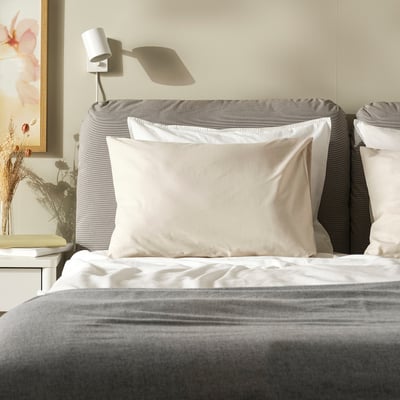 Taie d’oreiller crème DVALA, coton doux, forme carrée, sur un grand lit, avec tête de lit grise, design simple.
