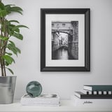 Photo encadrée en noir sur un mur blanc, un canal avec un pont et des objets décoratifs en dessous.