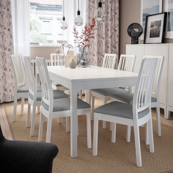 Salle à manger lumineuse avec chaises et table EKEDALEN blanches, rideaux légers, décor floral et ampoules suspendues.