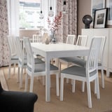 Salle à manger lumineuse avec chaises et table EKEDALEN blanches, rideaux légers, décor floral et ampoules suspendues.