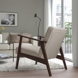 Salon moderne avec fauteuil beige EKENÄSET, cadre en bois, coussins moelleux, grande fenêtre adjacente et canapé confortable avec oreillers et une table.