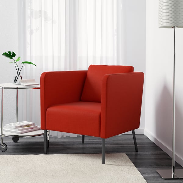 Un fauteuil rouge vif avec des pieds en métal, offrant un soutien lombaire et un coussin de dos réversible, positionné dans une pièce moderne avec une table d’appoint et une lampe.