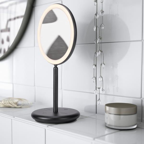 Un miroir de courtoisie moderne avec éclairage LED intégré se trouve sur un comptoir. Le miroir a une base ronde et un port USB pour la recharge.