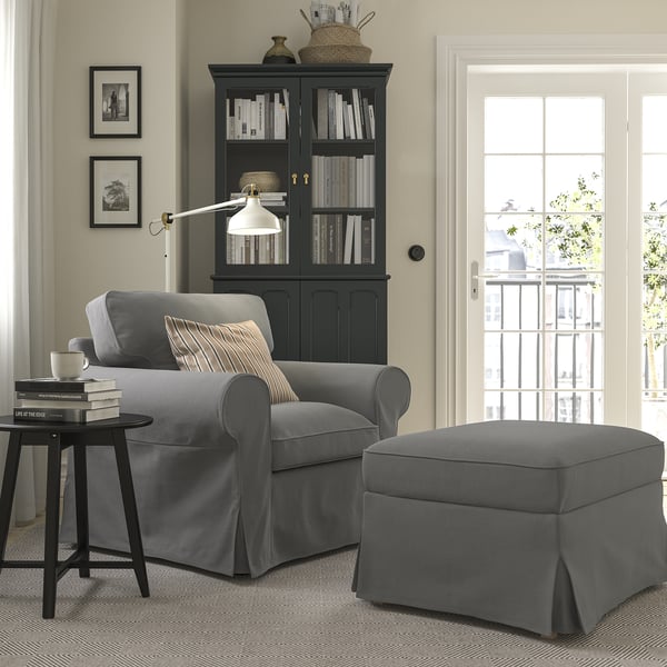 Salon confortable avec fauteuil gris EKTORP et pouf, armoire noire avec livres, lampe et œuvres d’art.