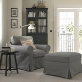 Salon confortable avec fauteuil gris EKTORP et pouf, armoire noire avec livres, lampe et œuvres d’art.