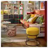 Salon vibrant : canapé orange avec coussins colorés, tabouret jaune, tapis rayé et étagères en bois avec décor.