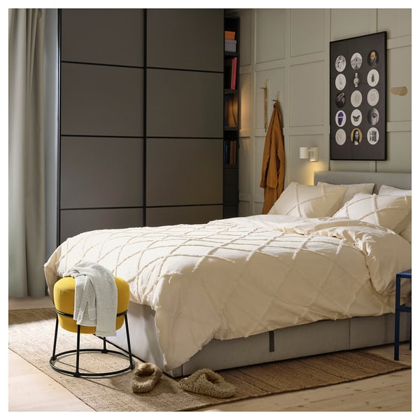 Chambre avec pouf EMMISLÖV : lit blanc avec couette à motifs, pouf moderne jaune, patère murale, revêtement en tissu.