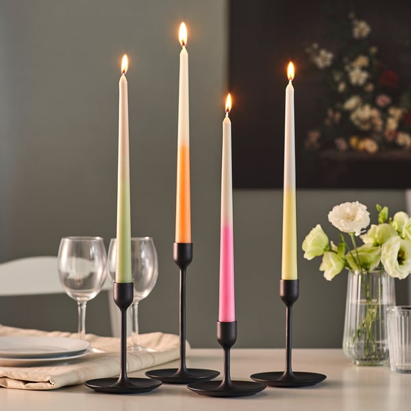 Quatre bougies coniques colorées dans des supports noirs sur une table à manger, éclairées et créant une atmosphère chaleureuse pour un dîner.