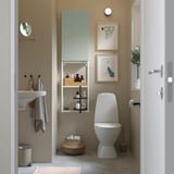 Salle de bain beige avec armoire gris-vert et étagères à côté des toilettes.