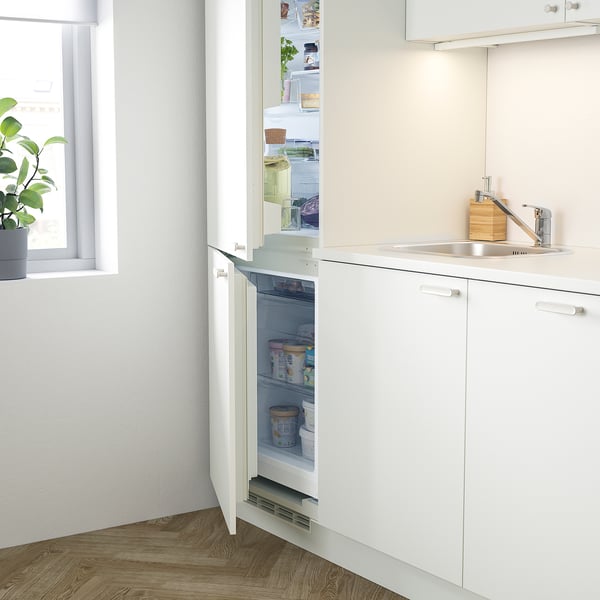 Cuisine minimaliste avec réfrigérateur intégré, évier et rangement. Armoires blanches, évier en acier inoxydable.