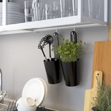 Des casseroles murales noires peuvent contenir des ustensiles de cuisine et des herbes fraîches. Des armoires blanches rangent les verres et les assiettes. Une planche à découper en bois s’appuie contre le mur.