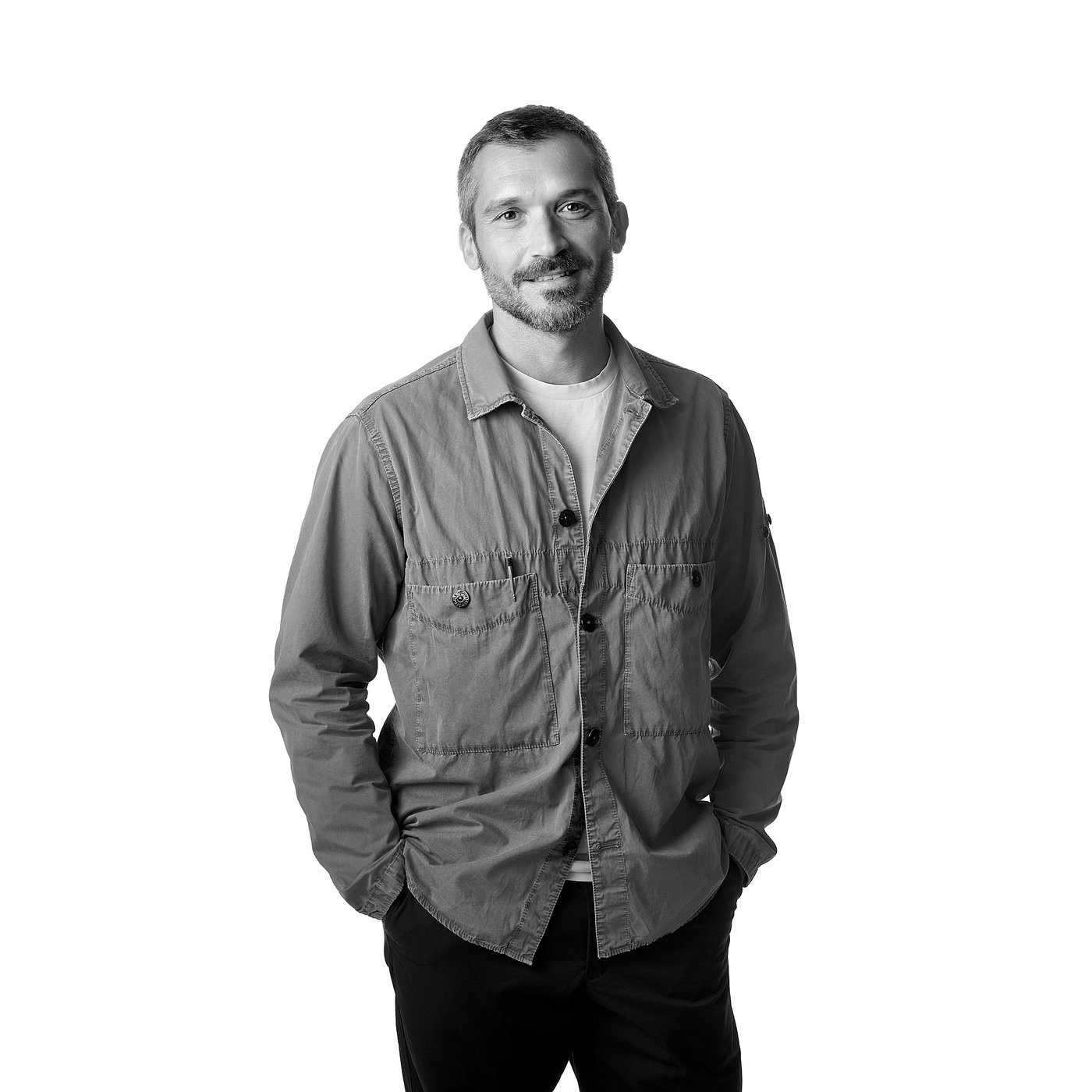 Portrait en noir et blanc de Gio Tirotto les mains dans les poches, vêtu d’une chemise à manches longues et d’un pantalon sombre.
