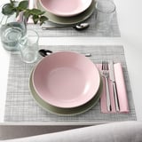 Deux couverts avec des bols roses et verts, de l’argenterie, des verres et des sets de table tissés sur une table blanche.