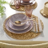 Set de table avec de la vaisselle en grès FÄRGKLAR dans des tons sourds, y compris des assiettes, un bol et du verre. Couverts dorés reposent sur un napperon tissé.