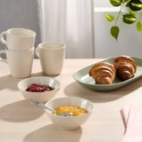 Une scène de cuisine lumineuse avec des bols FÄRGKLAR remplis de confiture et de fruits, accompagnés de tasses et de croissants.