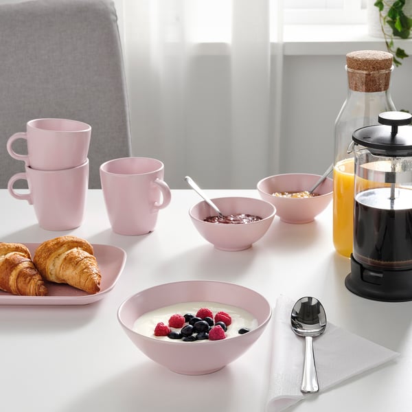 Un petit-déjeuner avec des bols en céramique rose, des tasses et une assiette avec des croissants sur une table blanche.