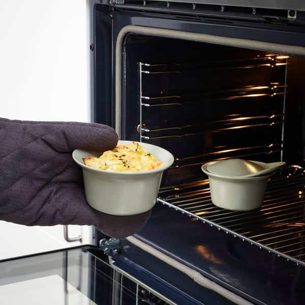 Une main tenant un petit plat en céramique avec des aliments cuits au four, prêts à être placés dans un four ouvert, à côté d’un autre plat similaire à l’intérieur.