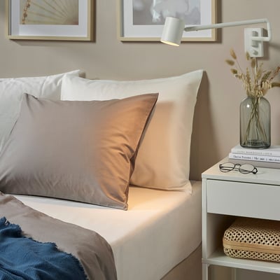 Chambre minimaliste avec literie beige FÄRGMÅRA sur un lit aux tons neutres, associée à une table d’appoint blanche exposant des fleurs séchées et des livres.