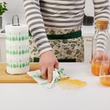 Une femme nettoie le comptoir avec du papier essuie-tout FAMILJ absorbant et recyclé avec des motifs de légumes verts. Jus renversé à proximité, tablier usé.