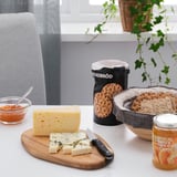 Planche à découper en bois avec fromage et couteau ; pot de miel, craquelins, confiture et plante.