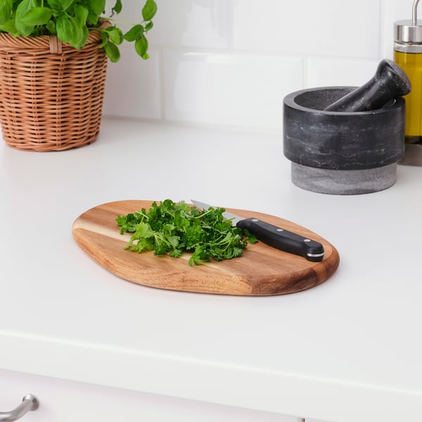 Planche à découper en bois avec couteau et herbes sur un comptoir blanc. Récipient de plante en osier et mortier en arrière-plan.