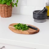 Planche à découper en bois avec couteau et herbes sur un comptoir blanc. Récipient de plante en osier et mortier en arrière-plan.