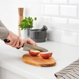 Personne tranche une pomme sur une planche à découper en bois d’acacia FASCINERA ; Comptoir de cuisine, dosseret de carreaux de métro blancs visibles.