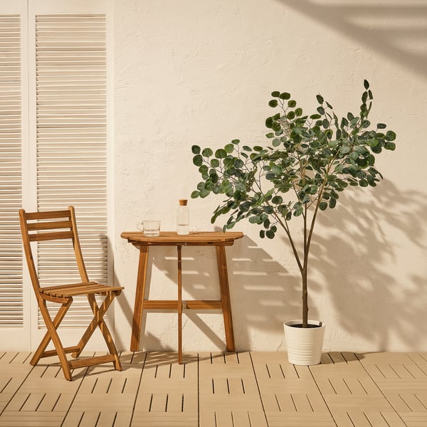 Petit ensemble de patio en bois avec une chaise pliante et une table, parfait pour la détente en plein air. À côté, une plante artificielle FEJKA en pot ajoute une beauté naturelle.