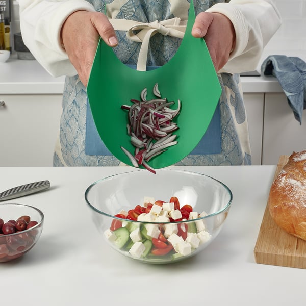 Personne tenant une planche à découper flexible avec des oignons émincés, les glissant dans un saladier contenant des tomates, des concombres et du fromage.